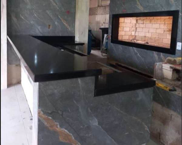 Bancada em pedra escura em área gourmet com churrasqueira ao fundo
